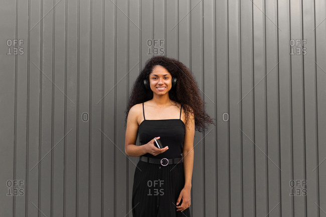 Positive ethnic female with Afro hairstyle standing in city and enjoying sound and songs in wireless headphones and browsing smartphone