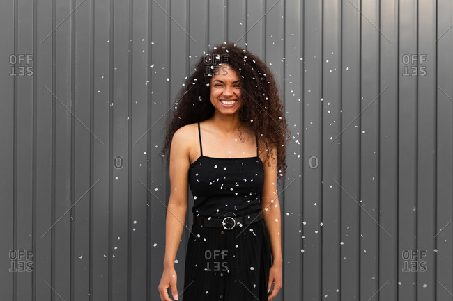 Joyful African American female with curly hair scattering white confetti in city while having fun and looking at camera