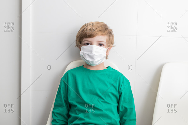 Calm boy in protective medical mask sitting on chair in medical room in clinic and looking at camera during COVID pandemic