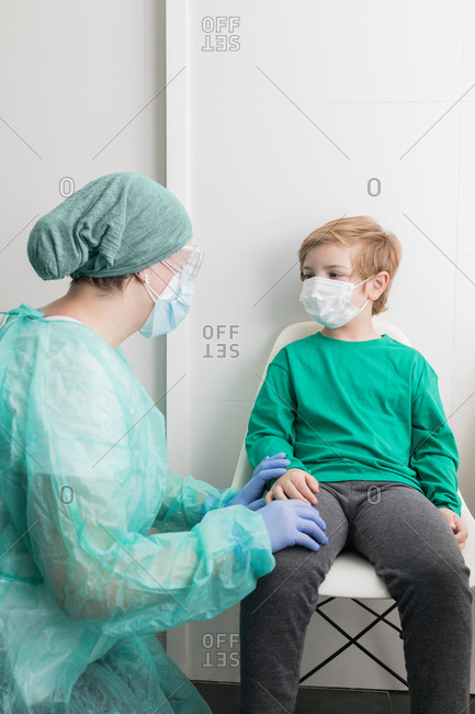 Physician in protective uniform speaking to kid in mask in medical office during coronavirus pandemic