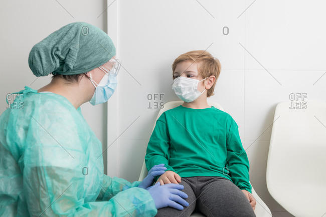 Physician in protective uniform speaking to kid in mask in medical office during coronavirus pandemic