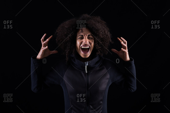 Stressful angry young African American female with curly hair shouting loudly and touching head while standing against black background