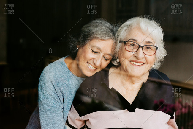 Through glass view of smiling mature woman gently embracing elderly mother looking at camera at home