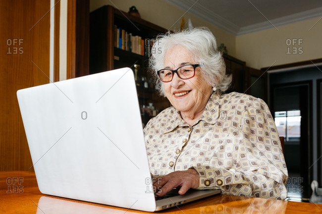 Positive senior female typing on modern netbook while sitting at table at home and reading news on social media