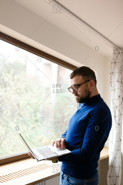 Young man with beard working on laptop by the window at home