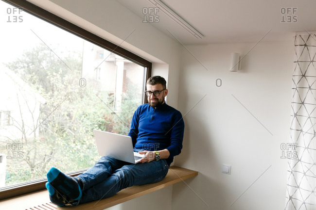 Young man with beard sitting on window ledge and working on laptop