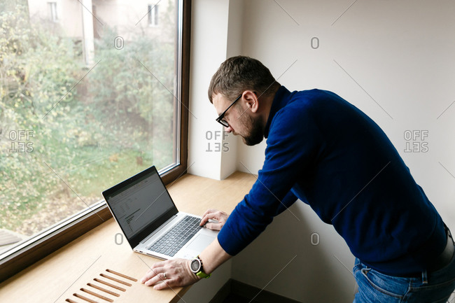 Young man with beard working on laptop by the window at home