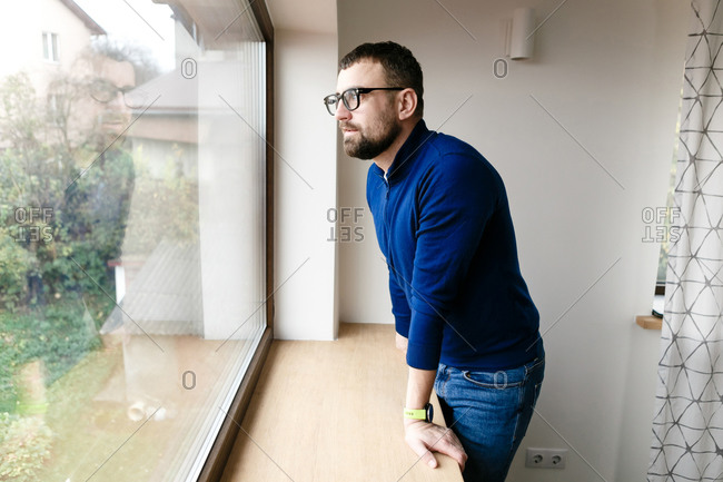 Young man with a beard in a blue jacket and jeans by the window at home