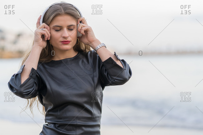 Pensive young woman wearing black dress walking along the seaside while listening to music on her headphones
