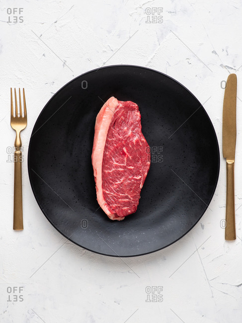 Raw beef black angus steak served on black ceramic plate with gold fork and knife over white background