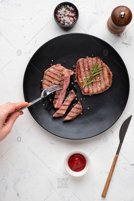 Top view of grilled beef steak served on black ceramic plate. Person cutting a piece of steak