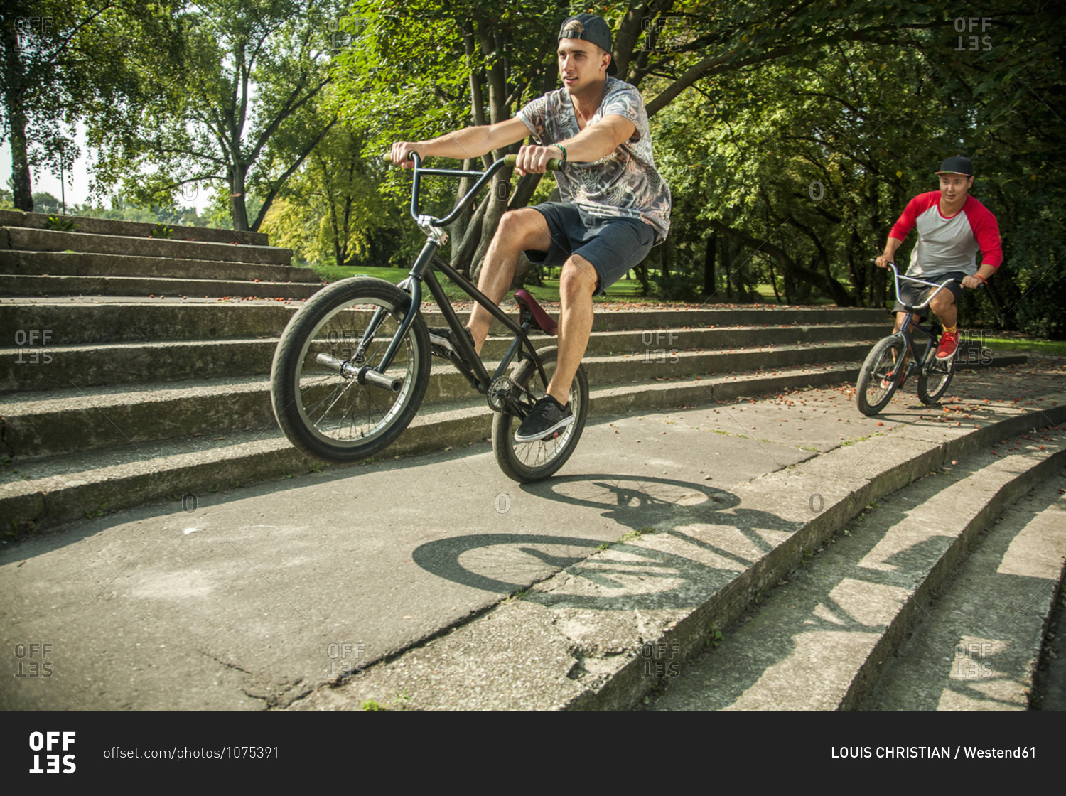 Young man doing wheelie stunt with BMX bicycle against friend cycling on steps at public park