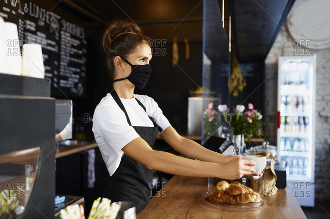 Female barista in protective face mask giving coffee while working at cafe