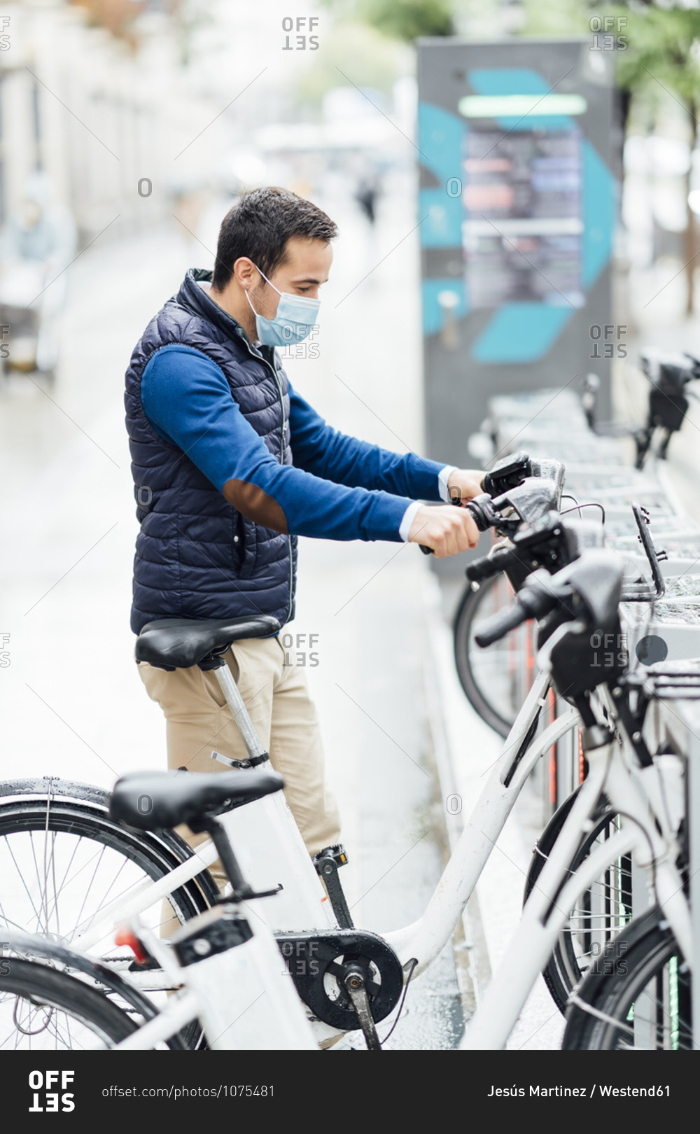 Young man wearing protective face mask holding electric bicycle at parking station during COVID-19