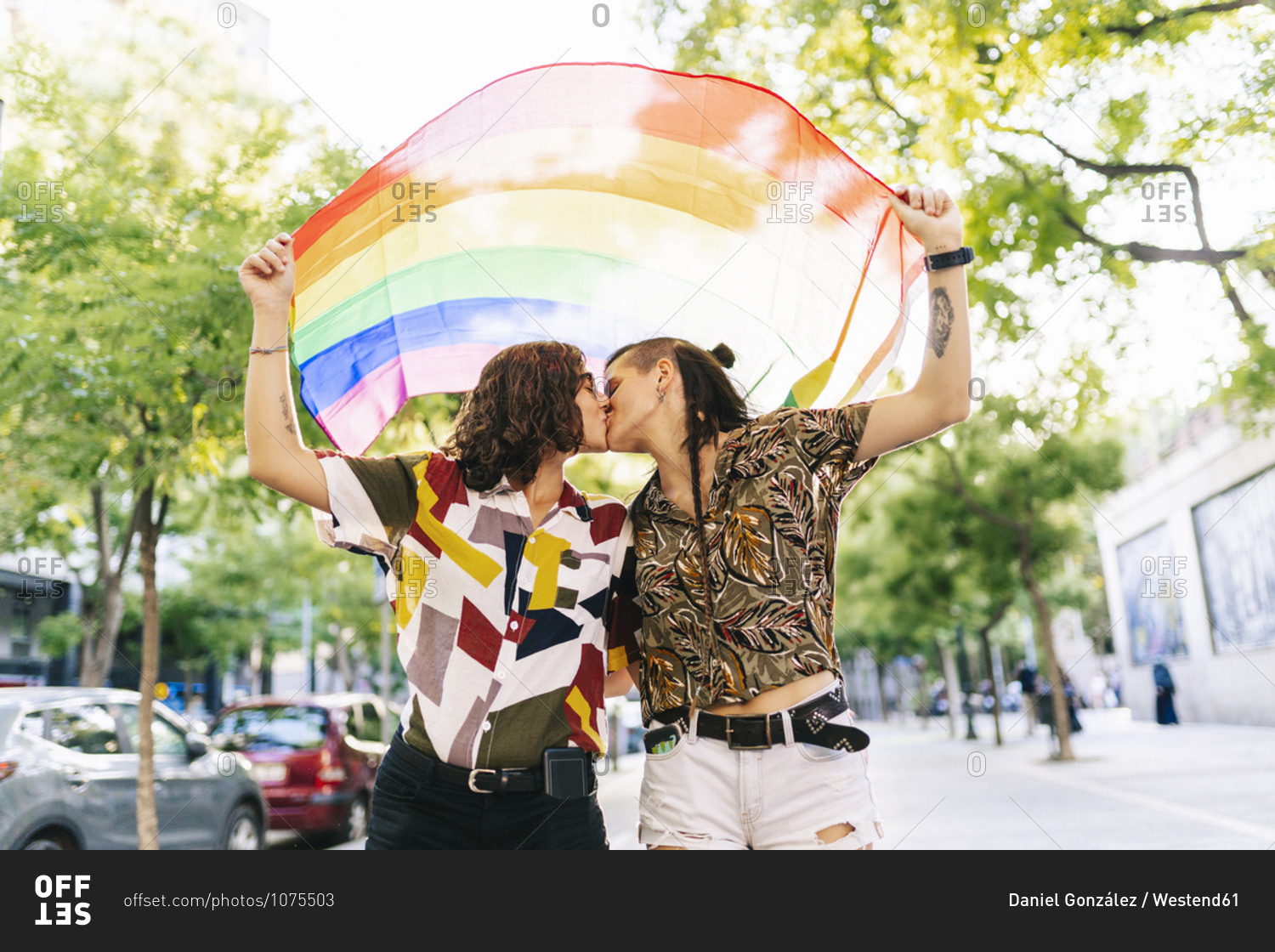 Lesbian Couple Holding Rainbow Flag While Kissing On Footpath In City