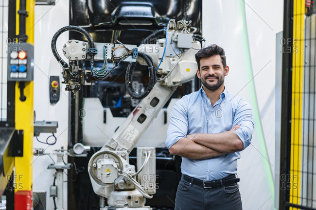 Smiling male entrepreneur with arms crossed standing against robotic arm at industry