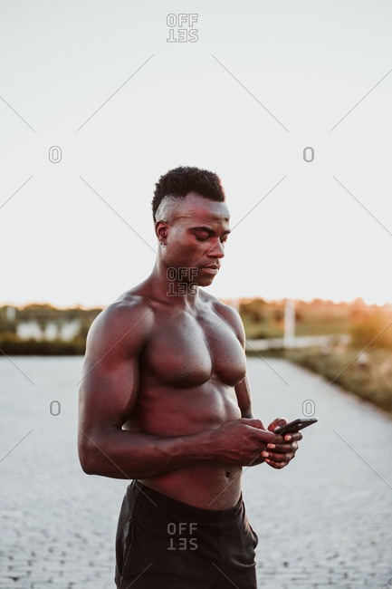 Young male athlete text messaging on smart phone while standing in park during sunset