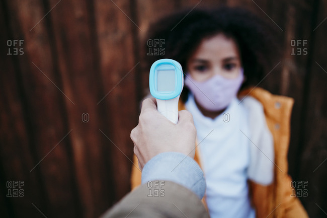 Woman using infrared thermometer while checking girl temperature against wooden wall