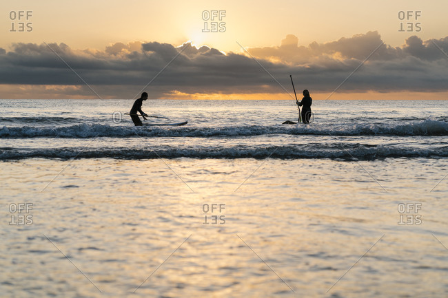 Friends surfing with paddleboard on sea against sky at dawn
