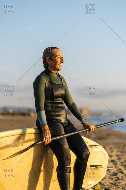 Smiling woman looking away while leaning on paddleboard at beach during dawn