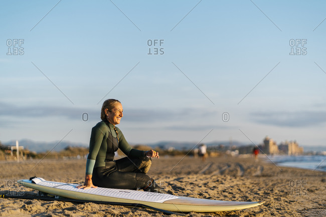 Smiling woman with paddleboard looking away while sitting on sand at beach during dawn