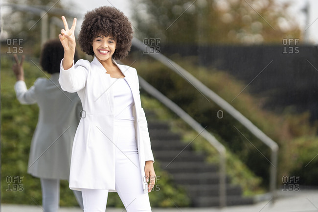Smiling businesswoman showing peace sign while standing against glass wall in city