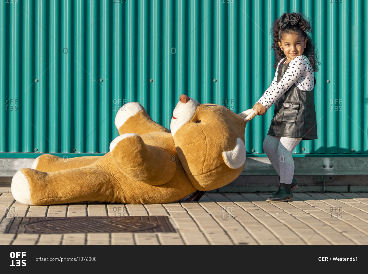 Girl dragging teddy bear on footpath while standing against green wall