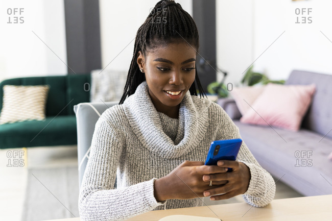 Smiling young woman text messaging on smart phone while sitting in living room at home
