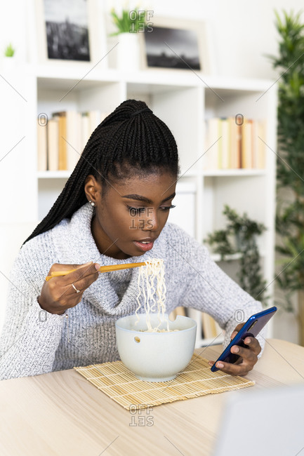 Young woman eating noodles text messaging on smart phone while sitting at home
