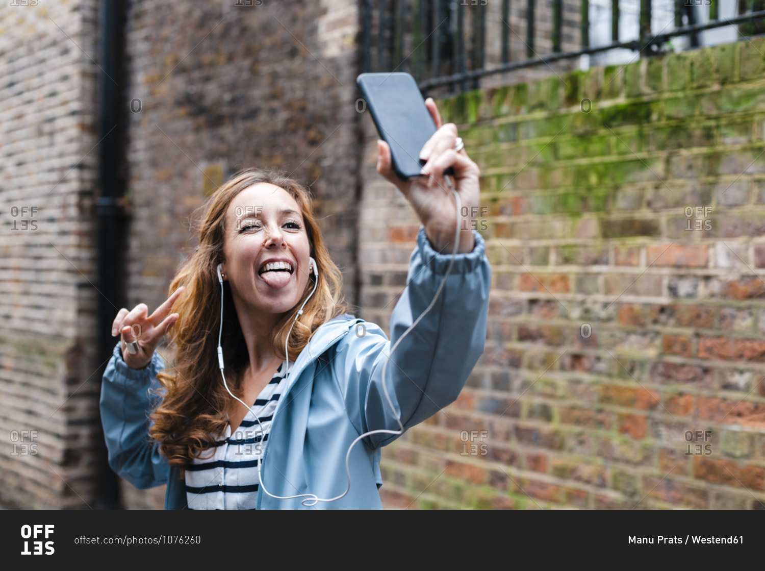 Carefree woman with in-ear headphones sticking tongue out while taking selfie through mobile phone in city