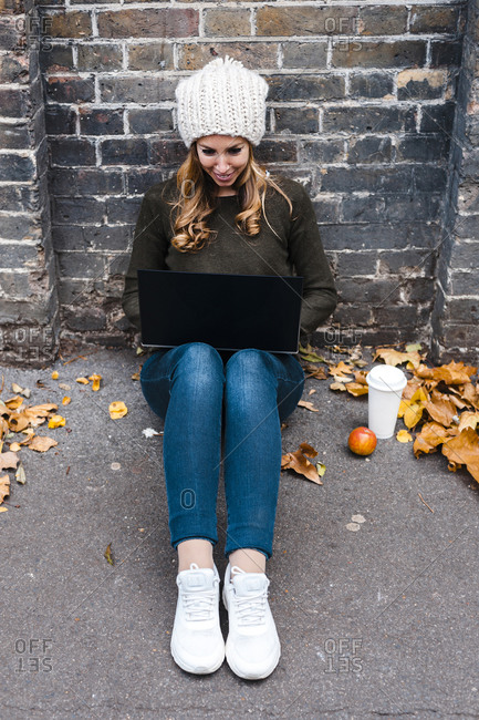 Woman using laptop while sitting against brick wall on footpath
