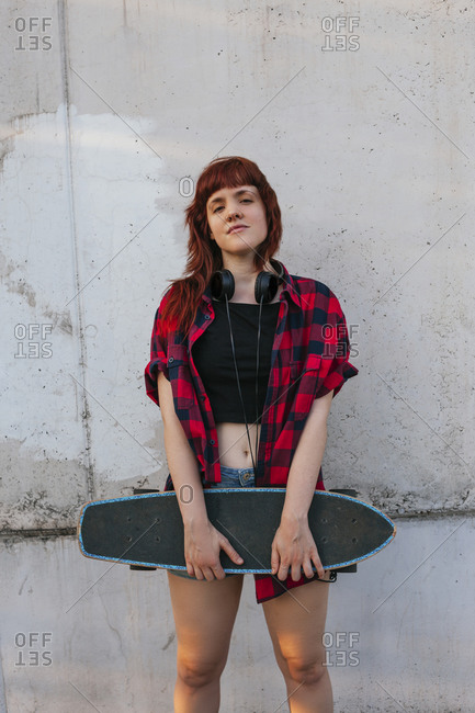 Young redhead woman with headphones holding skateboard against wall