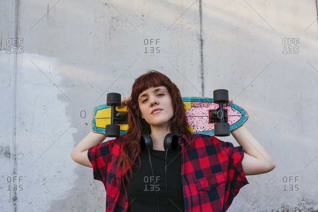 Redhead woman holding skateboard behind head against wall