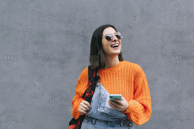 Smiling woman with sunglasses and purse using mobile phone while standing against gray wall