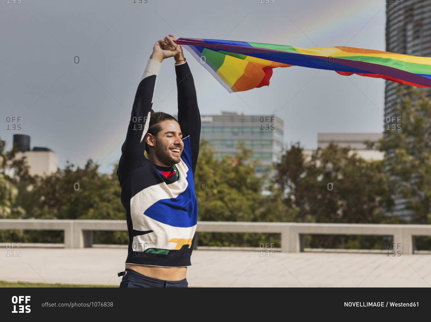 Smiling man with eyes closed holding rainbow scarf against sky