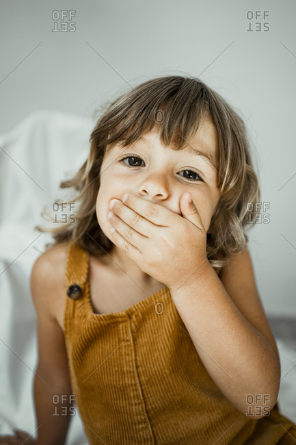 Cute girl covering mouth with hands while sitting against wall at home