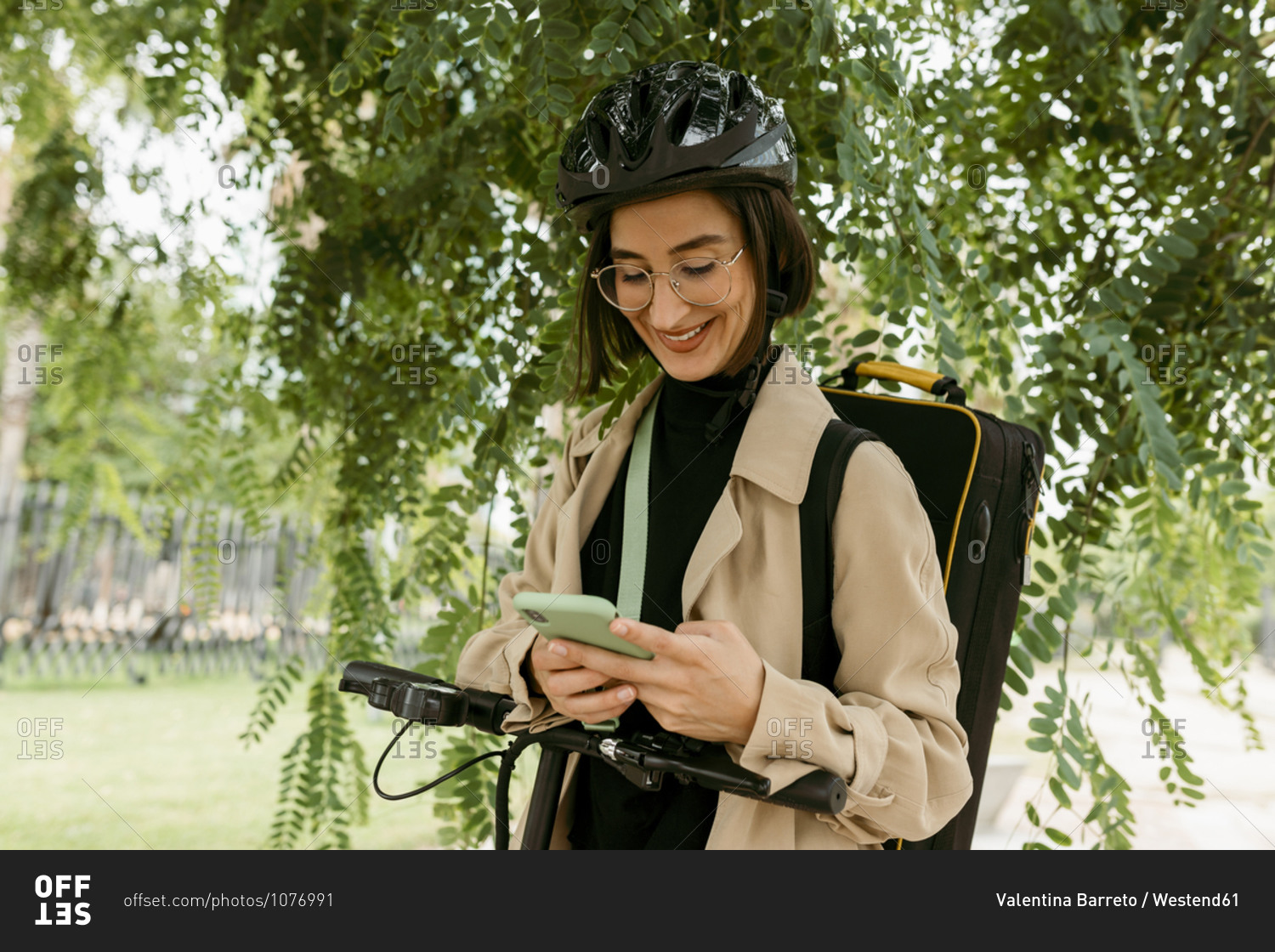 Smiling woman with instrument case and electric push scooter using mobile phone while standing under tree at park