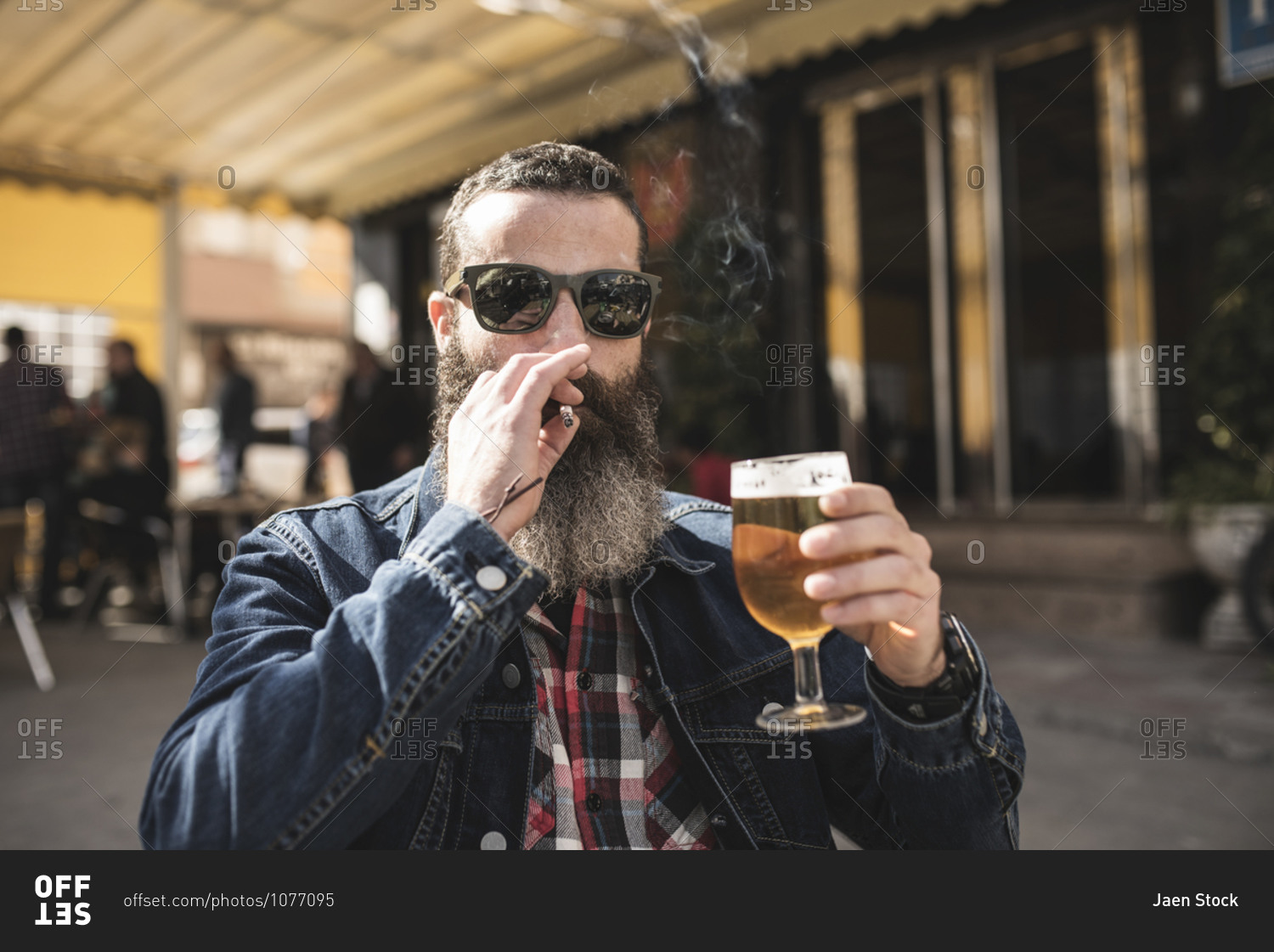 Bearded man smokes sitting on the terrace of a bar drinks a beer and looks at camera