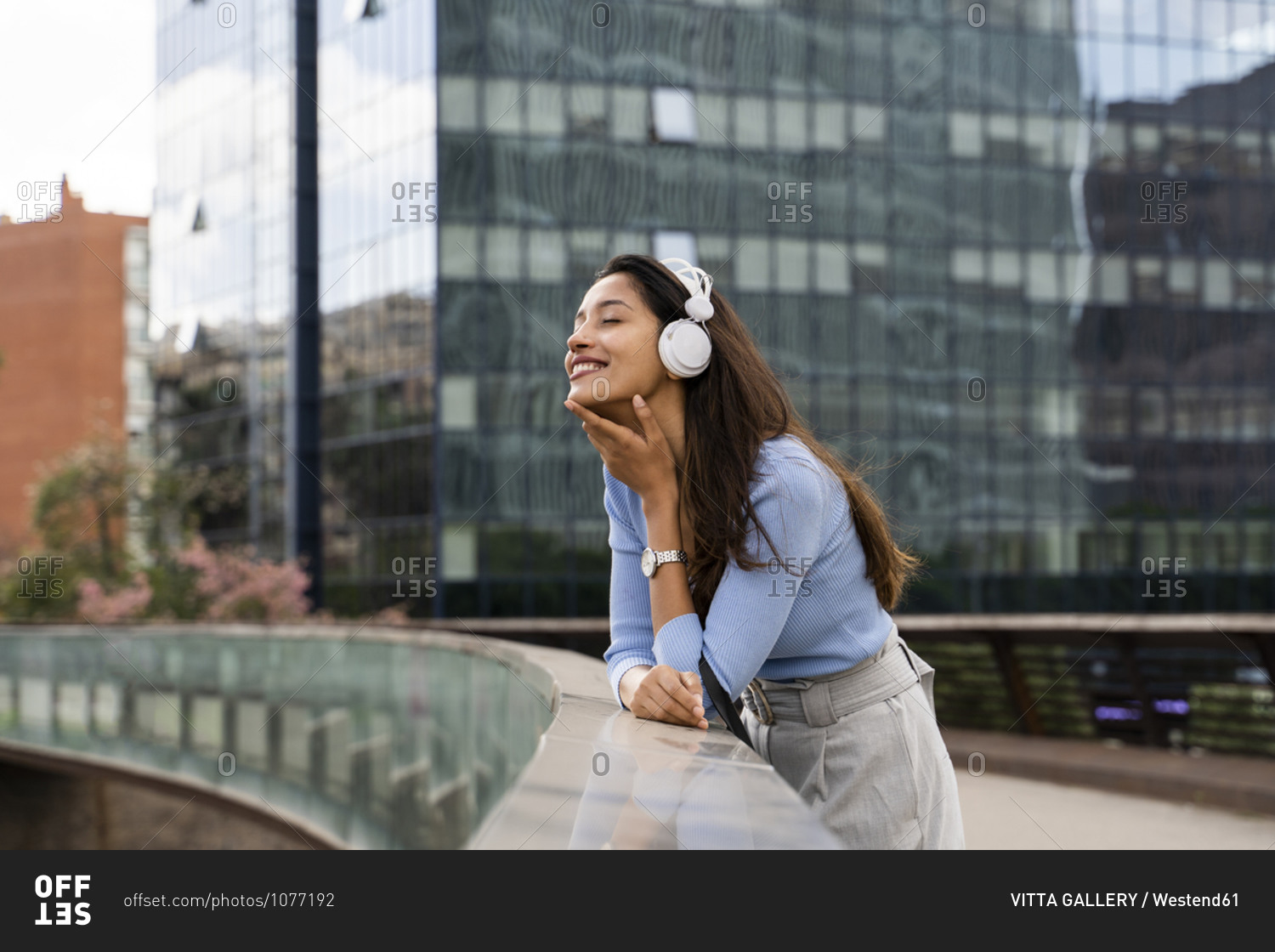Smiling businesswoman listening music through headphones while leaning on railing of footbridge