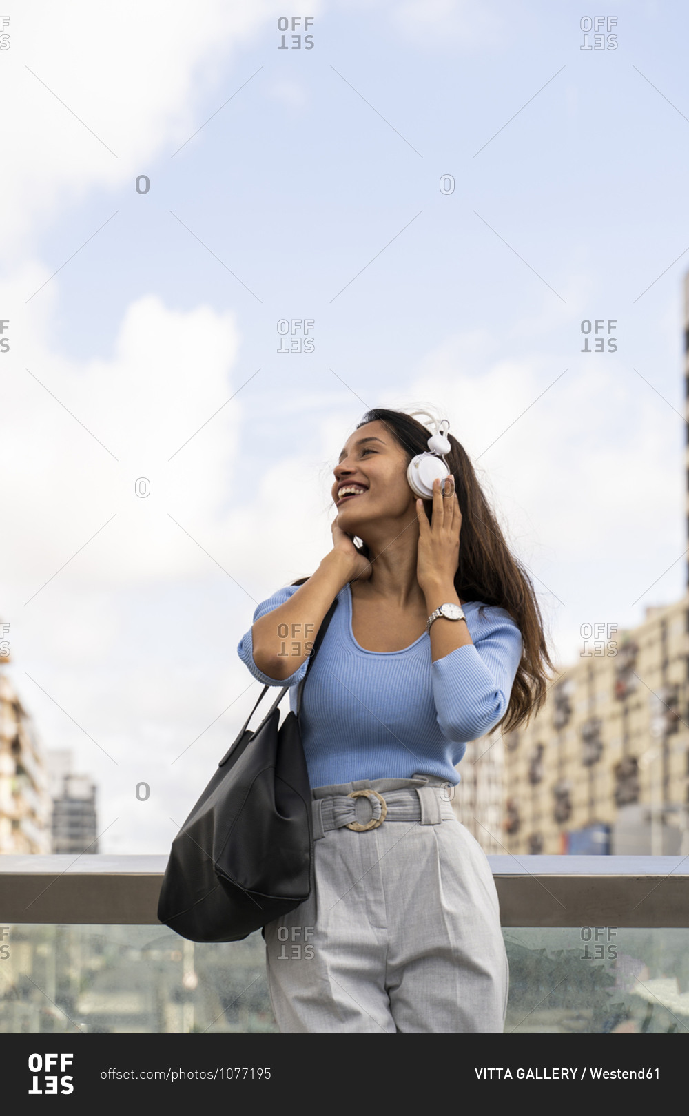 Happy businesswoman listening music through headphones while leaning on railing against sky