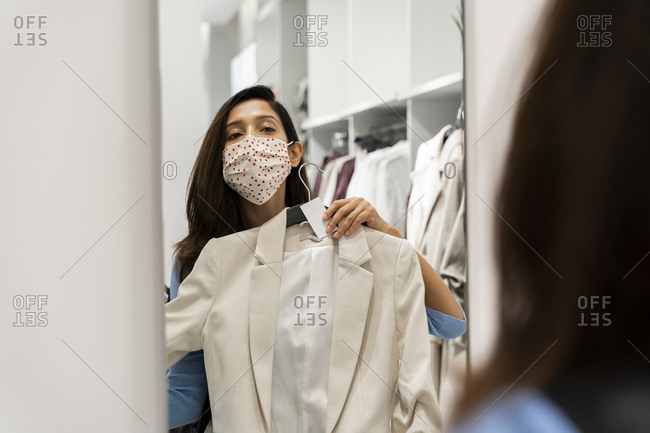 Woman wearing protective face mask while looking at reflection of suit in mirror at mall