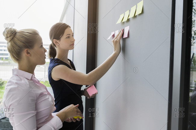 Female colleagues sticking adhesive notes on wall while discussing in office