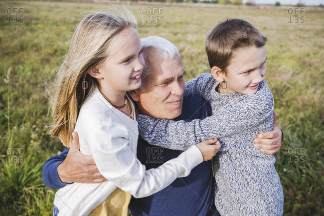 Grandfather looking away while embracing cute grandchildren on field
