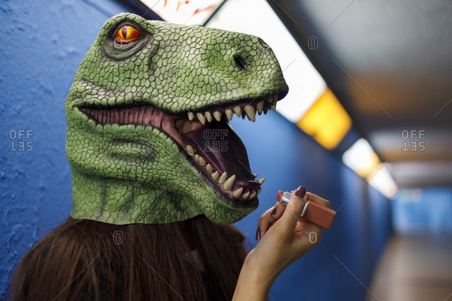 Woman applying lipstick while wearing dinosaur mask against blue wall