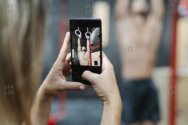 Close-up of young woman photographing boyfriend hanging on rings in gym