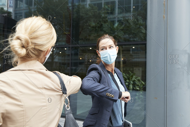Businesswoman and colleague with face mask greeting with elbow bump while standing against office building