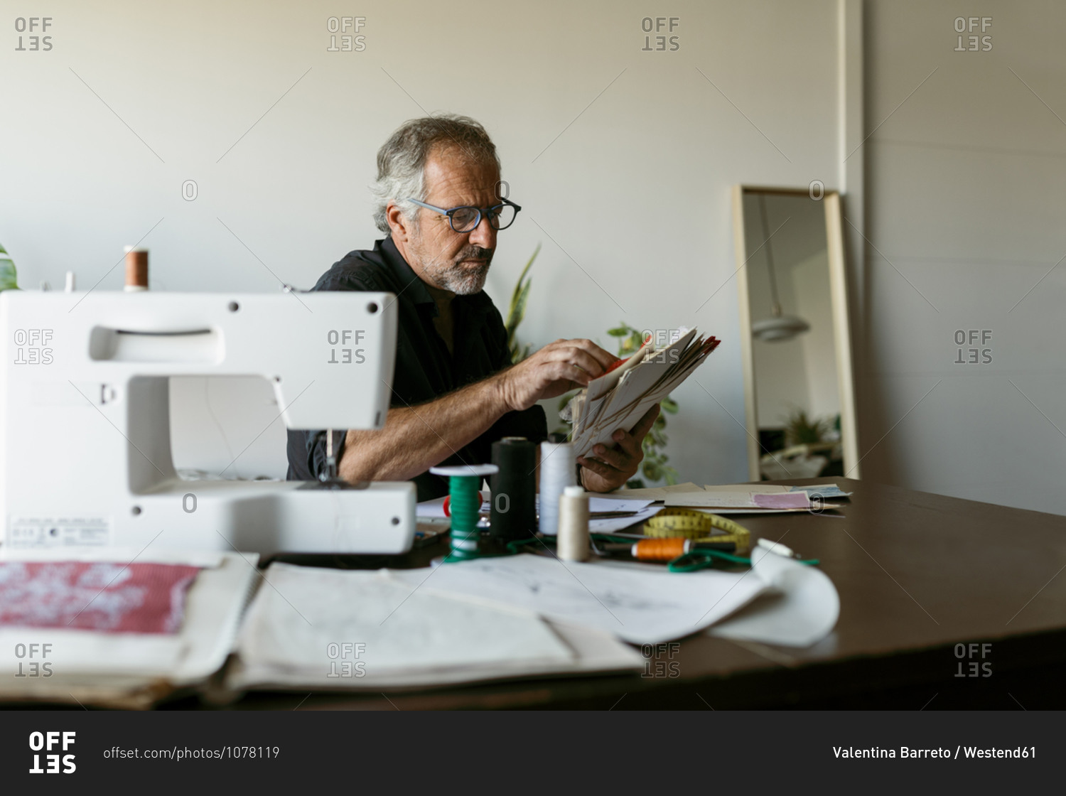 Mature tailor looking at fabric swatch papers while sitting at table in studio