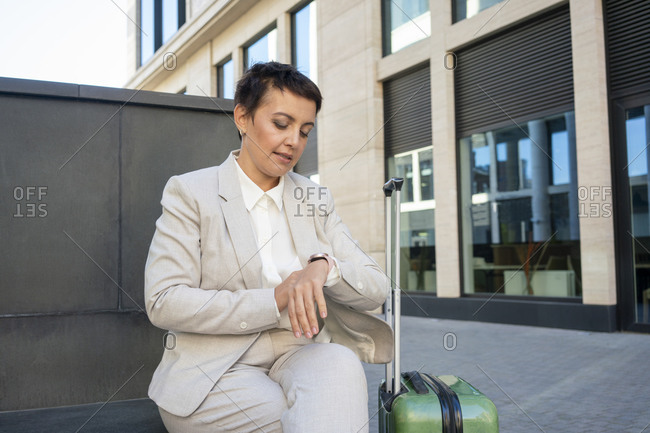 Businesswoman with suitcase checking time while sitting on bench against building