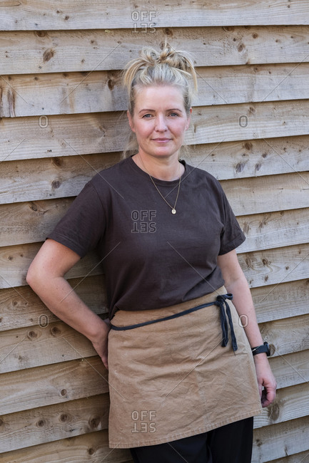 Portrait of waitress wearing brown apron, leaning against wall, smiling.