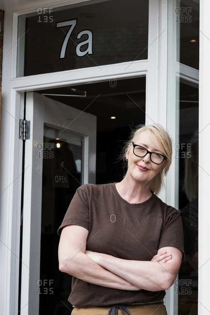Portrait of waitress with blond hair and glasses, leaning against entrance door, smiling at camera.